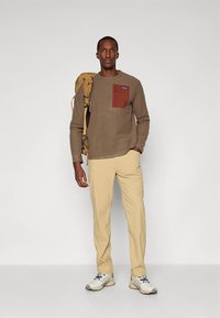 Maglione marrone con trama e tasca sul petto arancione, abbinato a pantaloni beige chiaro e sneakers bianche. Il modello porta uno zaino color senape.