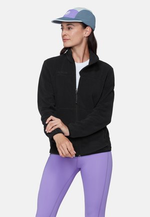 Žena v čiernej flisovej bunde na zips, levanduľových legínach a modro-levanduľovej šiltovke, ktorá sa pozerá doľava s prekríženými rukami.