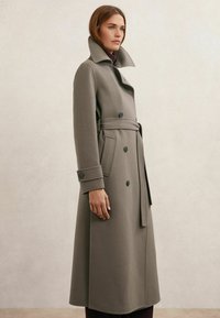 Dlhý sivý vlnený kabát s dvojradovým zapínaním, širokým golierom, opaskom v páse a manžetami na gombíky. Hladká textúra a priliehavý strih.