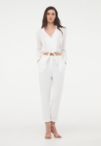 Camicia bianca a maniche lunghe con bottoni, cropped e con la parte anteriore annodata, abbinata a pantaloni bianchi a gamba larga con tasche laterali e vestibilità sartoriale.