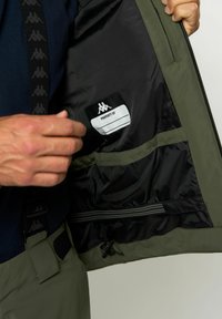 Zöld dzseki belső, amely egy fekete címkés zsebet mutat be "SZERZŐDÉS" felirattal, sima béléssel és állítható pánt részlettel.