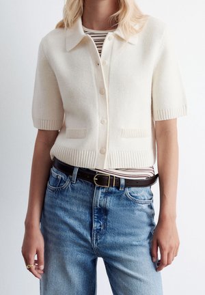 Femme portant un cardigan crème à manches courtes et boutonné par-dessus un top rayé, associé à un jean bleu et une ceinture noire avec une boucle dorée.