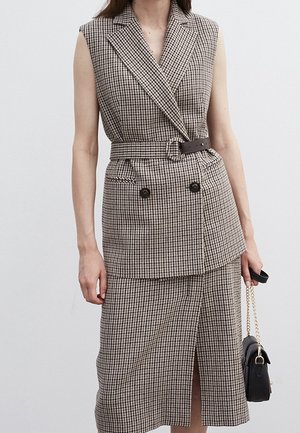 Veste sans manches ceinturée à motif pied-de-poule avec doubles boutons, associée à une jupe assortie longueur genou avec fente devant, tenant un sac à main noir.