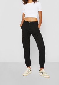 Pantaloni cargo neri con tasche laterali, polsini affusolati e chiusura con bottone, abbinati a una t-shirt bianca corta e sneaker beige.