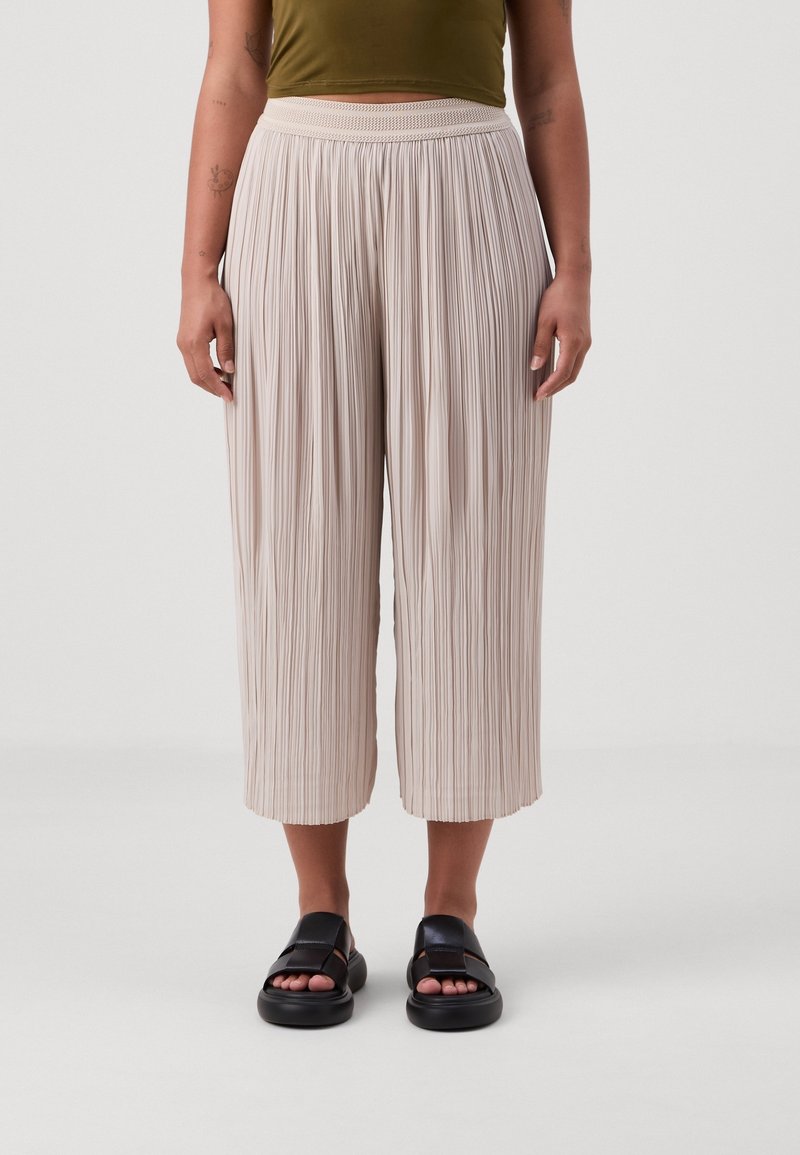 Pantalon plissé et court en tissu beige clair, avec une ceinture texturée. Porté avec des sandales noires, offrant une coupe décontractée et confortable.
