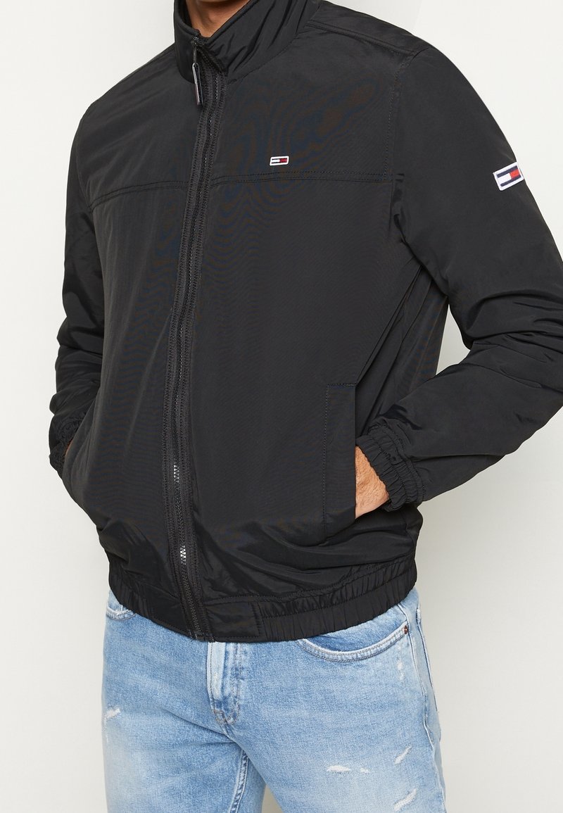 Giacca leggera nera con colletto alto, chiusura con zip e polsini elasticizzati. Presenta un piccolo logo sul lato sinistro del petto e sul braccio sinistro.