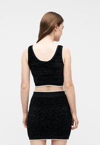 Γυναίκα με μακριά καστανά μαλλιά που φοράει μαύρο αμάνικο crop top και ψηλόμεση φούστα με λευκή διακόσμηση, στραμμένη προς τα πίσω, σε απλό φόντο.