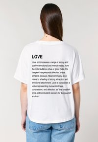Een persoon in een wit T-shirt met zwarte tekst die de liefde definieert, staande tegen een neutrale achtergrond. De achterkant toont het ontwerp van het shirt duidelijk.