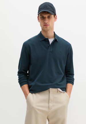 Mężczyzna w granatowej koszulce polo z długim rękawem, beżowych spodniach i czarnej czapce bejsbolowej, stojący z rękami w kieszeniach na jednolitym tle.