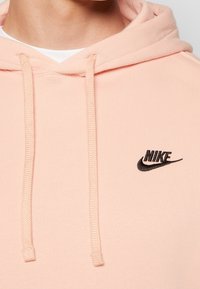 Rosa hoodie med en svart broderad Nike-logotyp på bröstet, med dragsnören och ett mjukt, texturerat tyg.