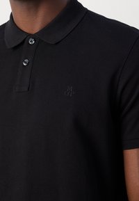 Polo noir à manches courtes avec deux boutons et un logo "MOP" brodé discrètement sur la poitrine gauche, porté par une personne.