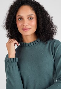 Teal gebreide trui met een volants halslijn en manchetten, voorzien van textuurrijke stippen details door de stof.