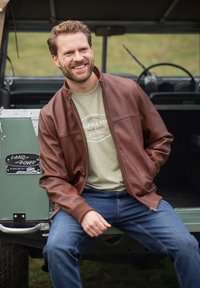 Smiling man wearing brown leather jacket, green graphic t-shirt, and blue jeans sits on the open back of a green Land Rover vehicle.