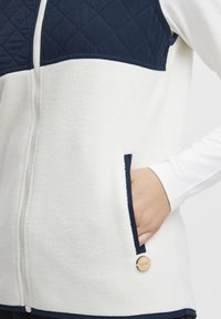 Bijela flis prsluk s tamnoplavim kviltiranim gornjim panelom, bočnim džepovima i kružnim drvenim detaljem s logotipom. Patent zatvarač se proteže duž prednje strane.
