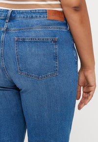 Jeans de denim azul con un diseño de corte recto, bolsillo trasero y una etiqueta de cuero marrón. Textura suave con un desvanecimiento mínimo.