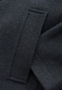 Dunkles Gewebe mit einer strukturierten Webart, ausgestattet mit einem flachen Panel und einer kleinen Klappe, hauptsächlich in Schwarz mit feinen blauen Untertönen.