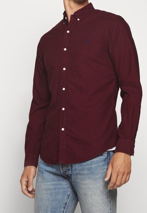 Mand iført en mørkerød skjorte med lange ærmer, hvide knapper og lyseblå jeans mod en ensfarvet baggrund.