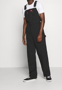 Salopette nera in canvas con bretelle regolabili, tasche frontali e un'etichetta rossa. Abbinata a una t-shirt bianca e sneakers nere.