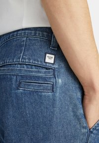 Jeans in denim blu con una tasca posteriore che presenta un'etichetta quadrata bianca con la scritta "GAS 1984." È visibile la cucitura lungo i bordi.