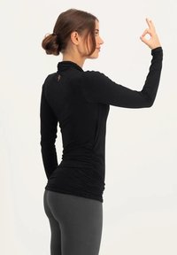 Schwarzes langärmliges Oberteil mit rüschenförmigem Design an der Taille, hohem Kragen und dezenter Logodetail auf der Rückseite, kombiniert mit grauen Leggings.