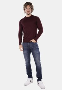 Camicia a maniche lunghe bordeaux abbinata a jeans blu. Il design è aderente e liscio, completato da sneakers bianche con accenti neri.