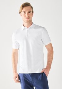 Camisa polo blanca de manga corta con un placket de dos botones. Tejido suave y liso; corte ajustado; combinada con pantalones azul marino.