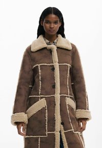 Cappotto in shearling marrone con rifiniture crema, ampio colletto e tasche a toppa. Presenta una chiusura con bottoni e un design a pannelli per un aspetto dai toni texture.