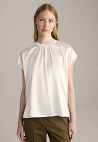 Lässige cremefarbene Bluse mit hohem Kragen, kurzen Ärmeln und gerafften Details am Kragen. Weicher, glatter Stoff.