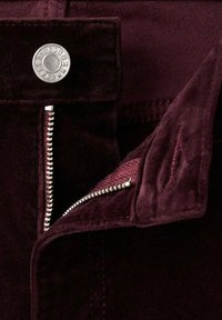 Pantalones de terciopelo burdeos con un botón plateado y cierre con cremallera. Las costuras visibles y una textura suave acentúan el diseño.