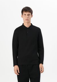 Un tânăr stă în picioare, purtând o cămașă polo cu mânecă lungă de culoare neagră și pantaloni negri, pe un fundal alb simplu.