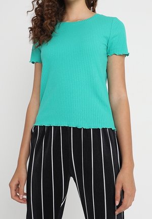 Kvinde iført en turkis ribstrikket kortærmet top med bølgede kanter og sorte bukser med lodrette hvide striber.