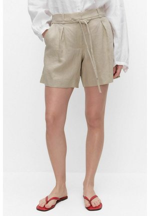 Femme portant un short en lin beige avec ceinture à nouer, un chemisier blanc à manches longues et des sandales tongs rouges, debout sur fond blanc.