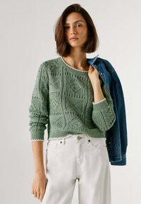 Ženska s rjavimi lasmi do ramen, oblečena v zelen pulover iz pletenine in bele hlače, drži modro jeans jakno prek enega ramena.