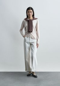 Γυναίκα στέκεται μπροστά σε απλό φόντο, φορώντας ένα ανοιχτό ροζ ζακέτα, λευκό παντελόνι με φαρδιά πόδια, μπεζ κάλτσες, μαύρα φλατ παπούτσια και ένα καφέ πουλόβερ περασμένο στους ώμους.