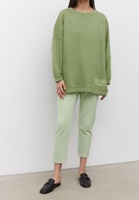 Felpa verde oversize con maniche lunghe, presenta una tasca applicata e dettagli testuali. Abbinata a pantaloni verde chiaro e scarpe slip-on nere.