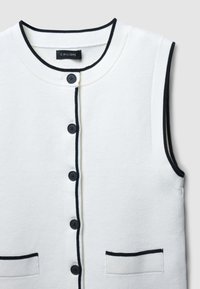 Weißes ärmelloses Oberteil mit schwarzer Borte, runder Ausschnitt, sechs schwarze Knöpfe vorne und zwei aufgesetzte Taschen mit schwarzen Akzenten.