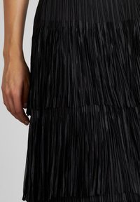 Falda negra plisada con flecos verticales, una textura suave y un diseño en capas. Mano visible sosteniendo la tela.