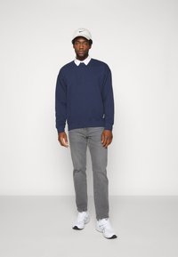 Marineblauwe sweatshirt met een wit collared shirt eronder, grijze jeans en witte sneakers, gedragen met een witte pet. Eenvoudig ontwerp.