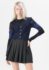 Jonge vrouw met lang blond haar, gekleed in een blauw en zwart langemouwtop, een donkerblauwe knoopvest en een zwarte geplooide rok.