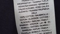 Etichetă de material pe care este scris „100% bumbac” în mai multe limbi pe un material alb pe fundal de țesătură închisă la culoare.
