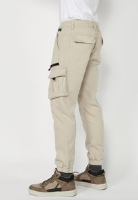 Beige Cargo-Hosen mit elastischen Bündchen und mehreren Taschen, ausgestattet mit schwarzen Reißverschlüssen und kontrastierenden Nähten. Getragen mit braunen High-Top-Sneakern.