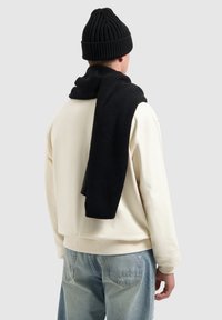 Zwarte gebreide beanie en sjaal over een lichtgekleurde sweatshirt, met ribbelstructuur en een relaxte pasvorm. Lichtblauwe jeans zichtbaar.