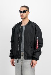 Black bomber jacket made of smooth fabric, featuring a quilted lining, ribbed cuffs, side pockets, and a red tag accent on the sleeve.