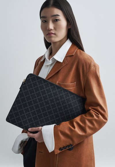 Čierny trojuholníkový laptop obal s textúrovaným povrchom, ktorý drží model v hnedom semišovom blejzri s kontrastnými bielymi manžetami.