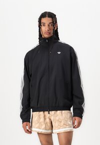 Črna zip-up jakna z belimi črtami na rokavih, z visokim ovratnikom in logotipom. Nosijo jo preko vzorčastih bež hlač.