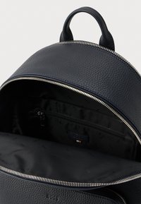 Juodas tekstūruotas odinis kuprinės krepšys su atvira pagrindine skyriaus dalimi, parodančia vidinį užtrauktuku uždaromą kišenių ir firminę minkštą pamušalą.