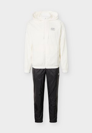 Chaqueta blanca con capucha y cremallera, con el logo EA7 en el pecho, combinada con pantalones deportivos negros texturizados.