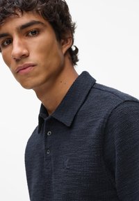 Granatowa koszulka polo z teksturowanej tkaniny, z krótkim kołnierzykiem i zapięciem na trzy guziki. Posiada wyszywane logo na piersi oraz subtelne akcenty haftu.