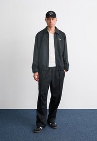 Black lightweight jacket with elastic cuffs, paired with loose-fitting black pants and black sneakers featuring white accents. Black baseball cap.