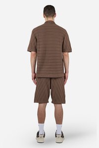 Brun skjorte med korte ermer og matchende shorts med teksturerte linjer. Skjorten har en enkel krage; shortsen har en rett snitt. Hvite sneakers er synlige.